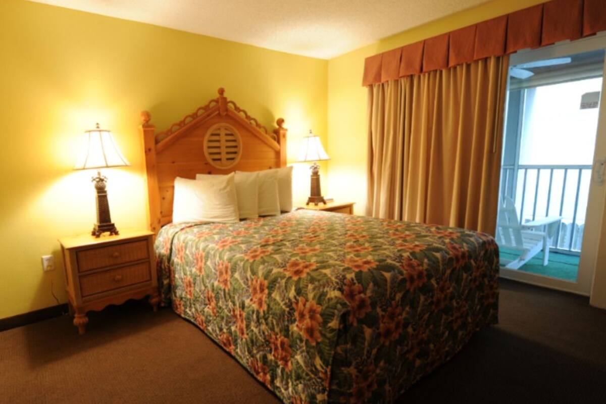 A hotel room with a floral bedspread, wooden headboard, two bedside lamps, a nightstand, and a view of the outside balcony.