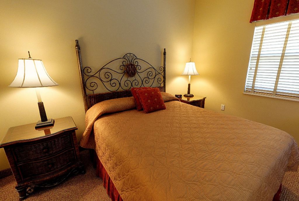A bedroom with a large bed, two bedside tables with lamps, a decorative headboard, and a window with blinds.