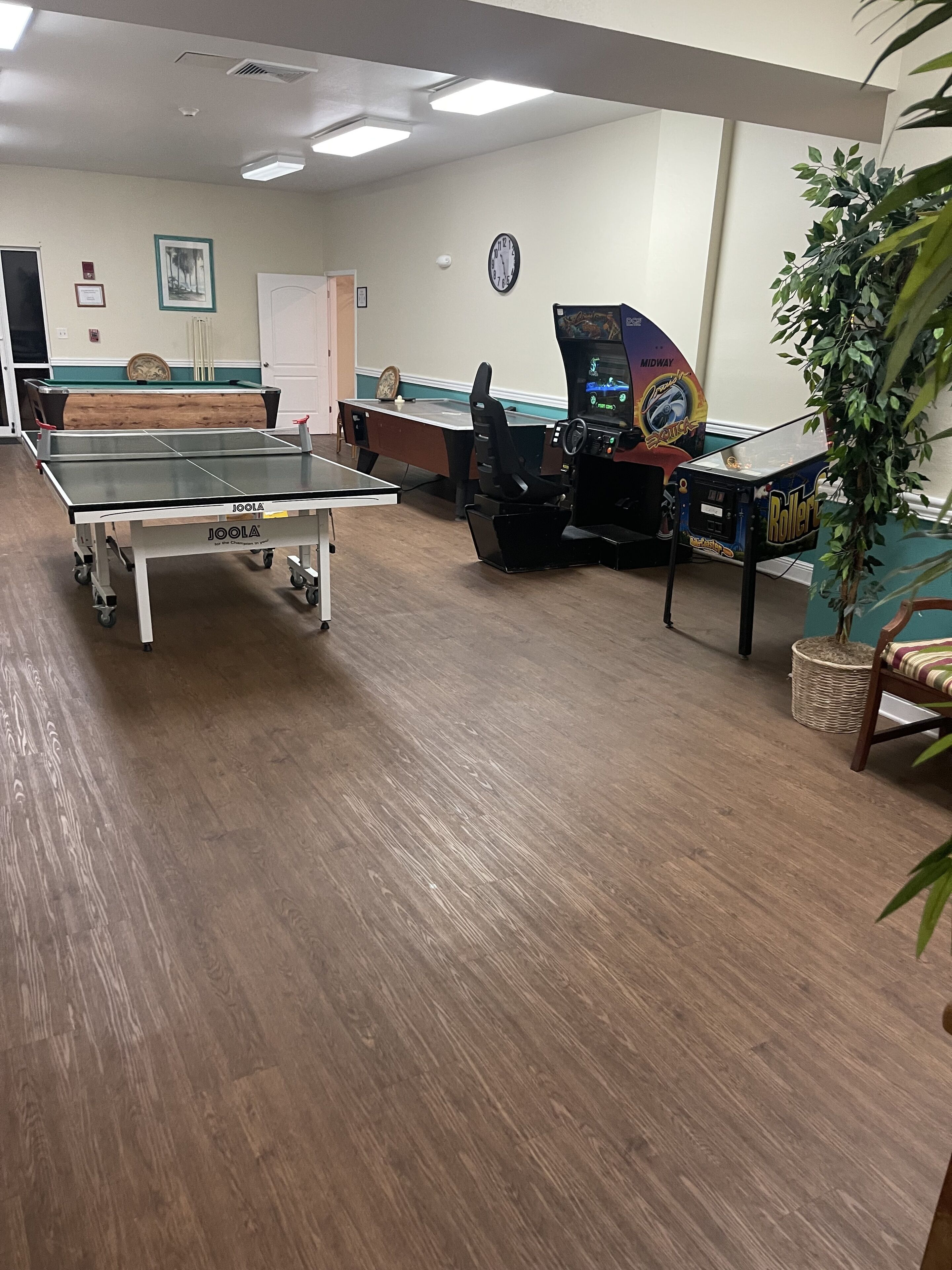 A billiards table, a pool table, and a pinball machine in a room with wooden flooring and a clock on the wall.