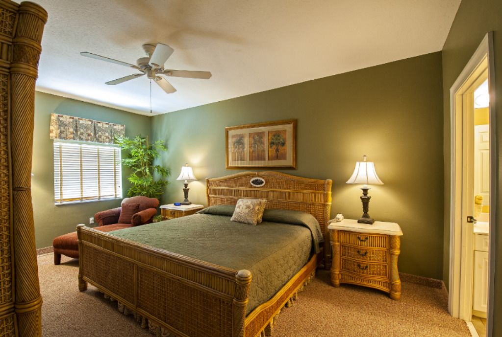 A bedroom with a large bed, two bedside tables, a nightstand, a chair, a ceiling fan, a window with blinds, and a door.