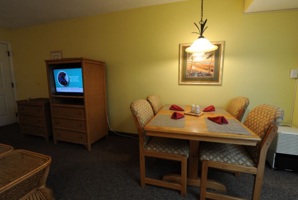 A dining area with a wooden table and chairs, a television on a wooden cabinet, and a framed picture on the wall.