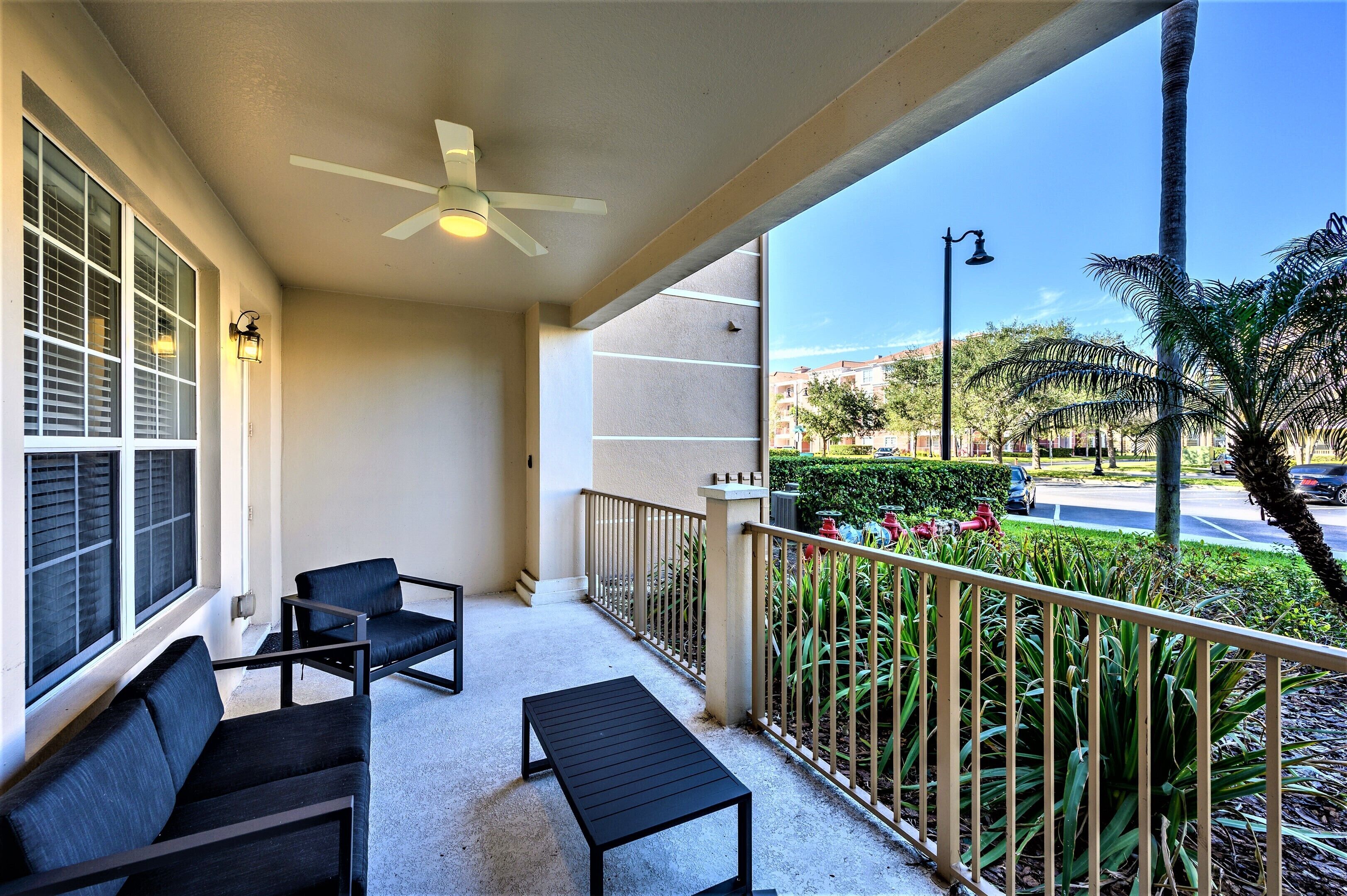 A balcony with two black chairs and a small table, a ceiling fan, and a view of a street with palm trees and buildings.