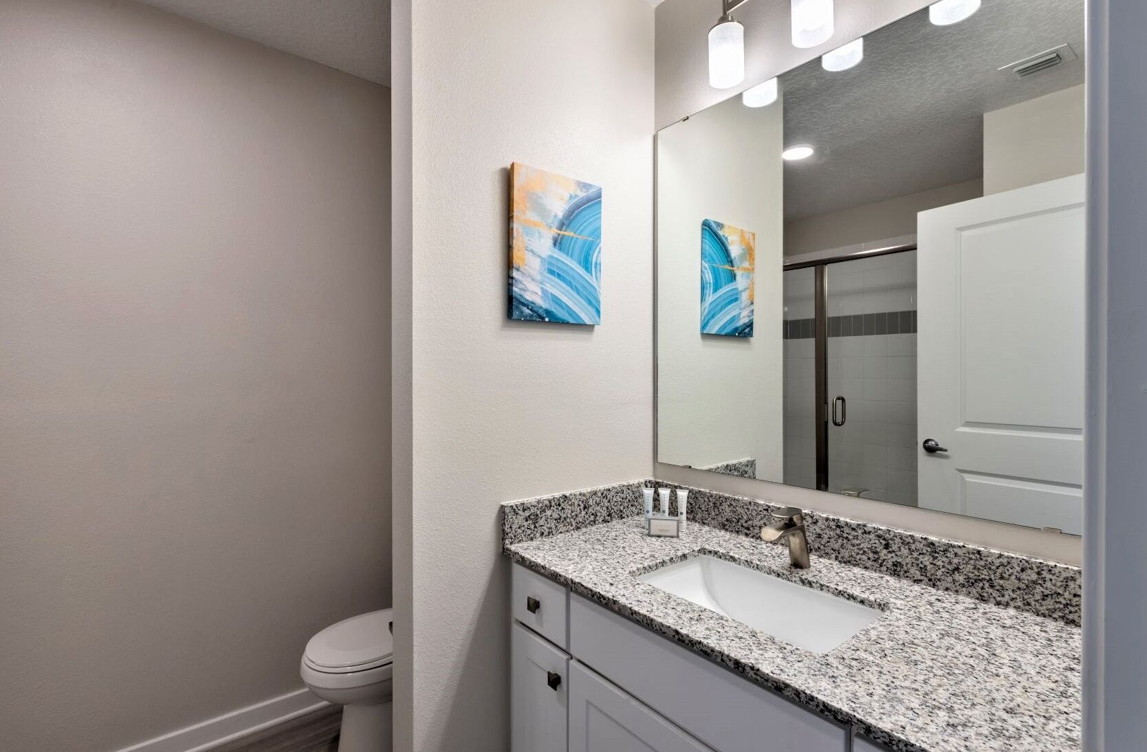 Un baño con lavamanos de granito, un espejo y dos cuadros abstractos en la pared.