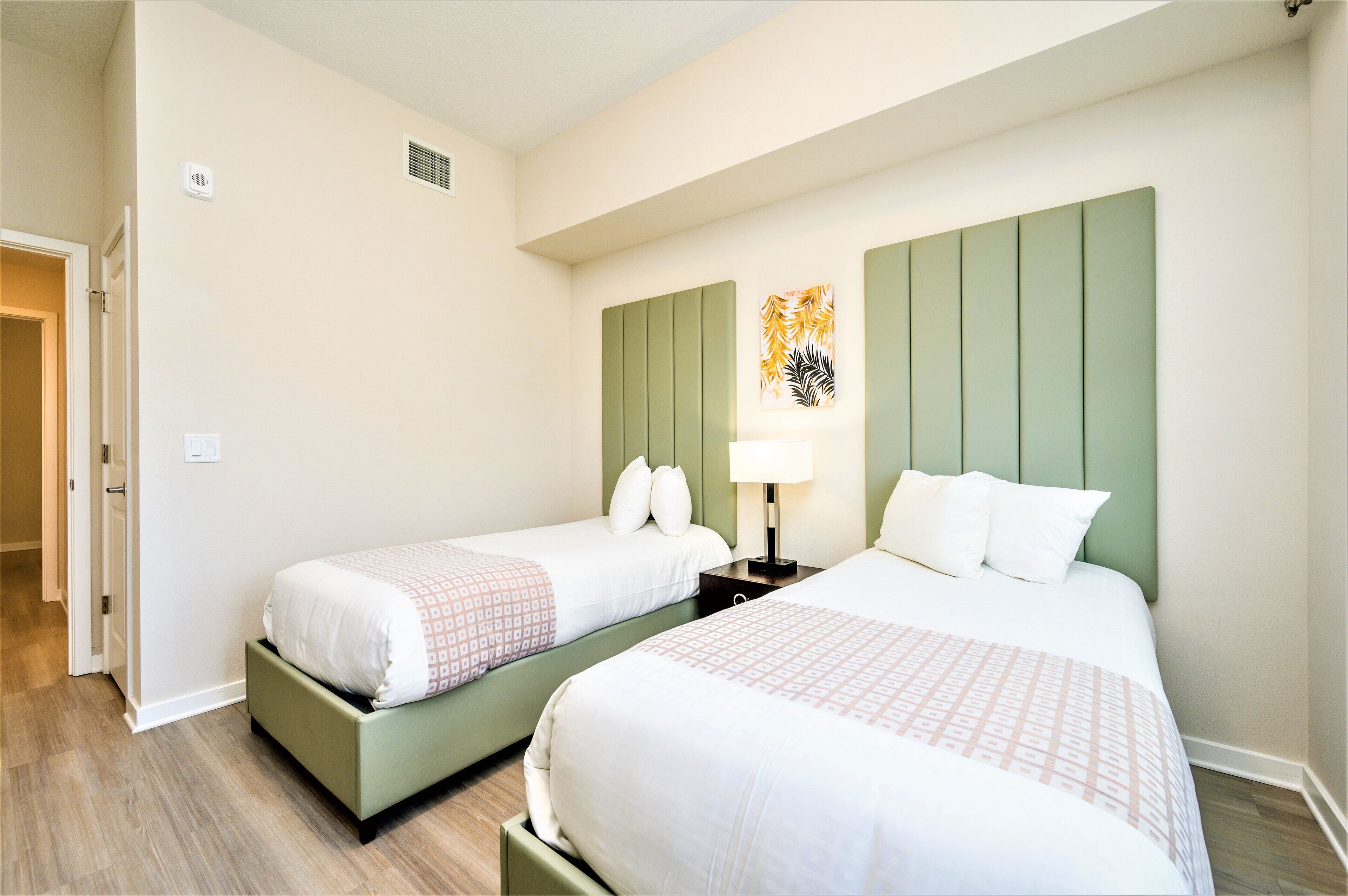 Une chambre d’hôtel avec deux lits, une tête de lit verte, une œuvre d’art accrochée au mur et une table de chevet avec une lampe.