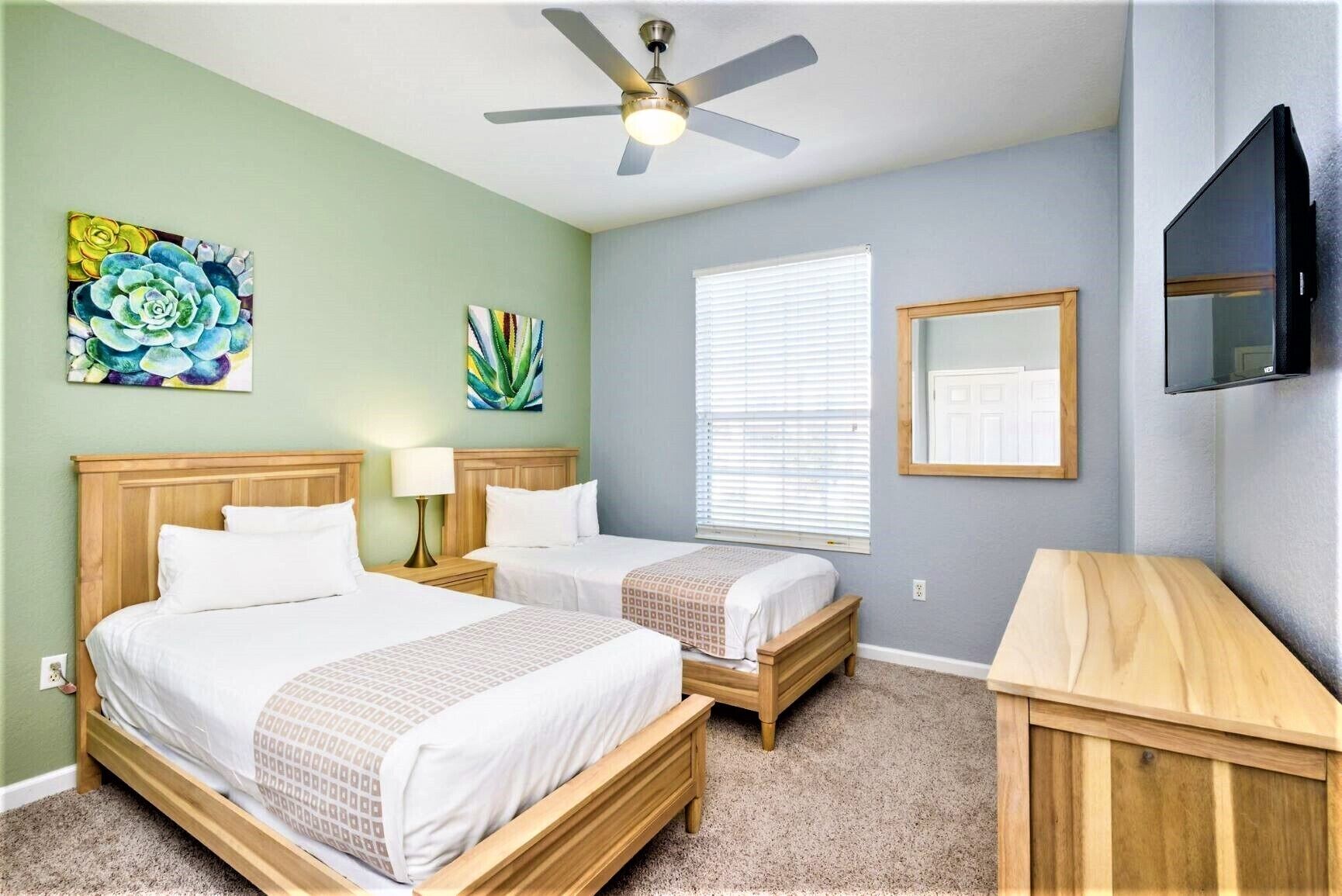 A hotel room with two beds, a ceiling fan, a TV, and artwork on the walls.