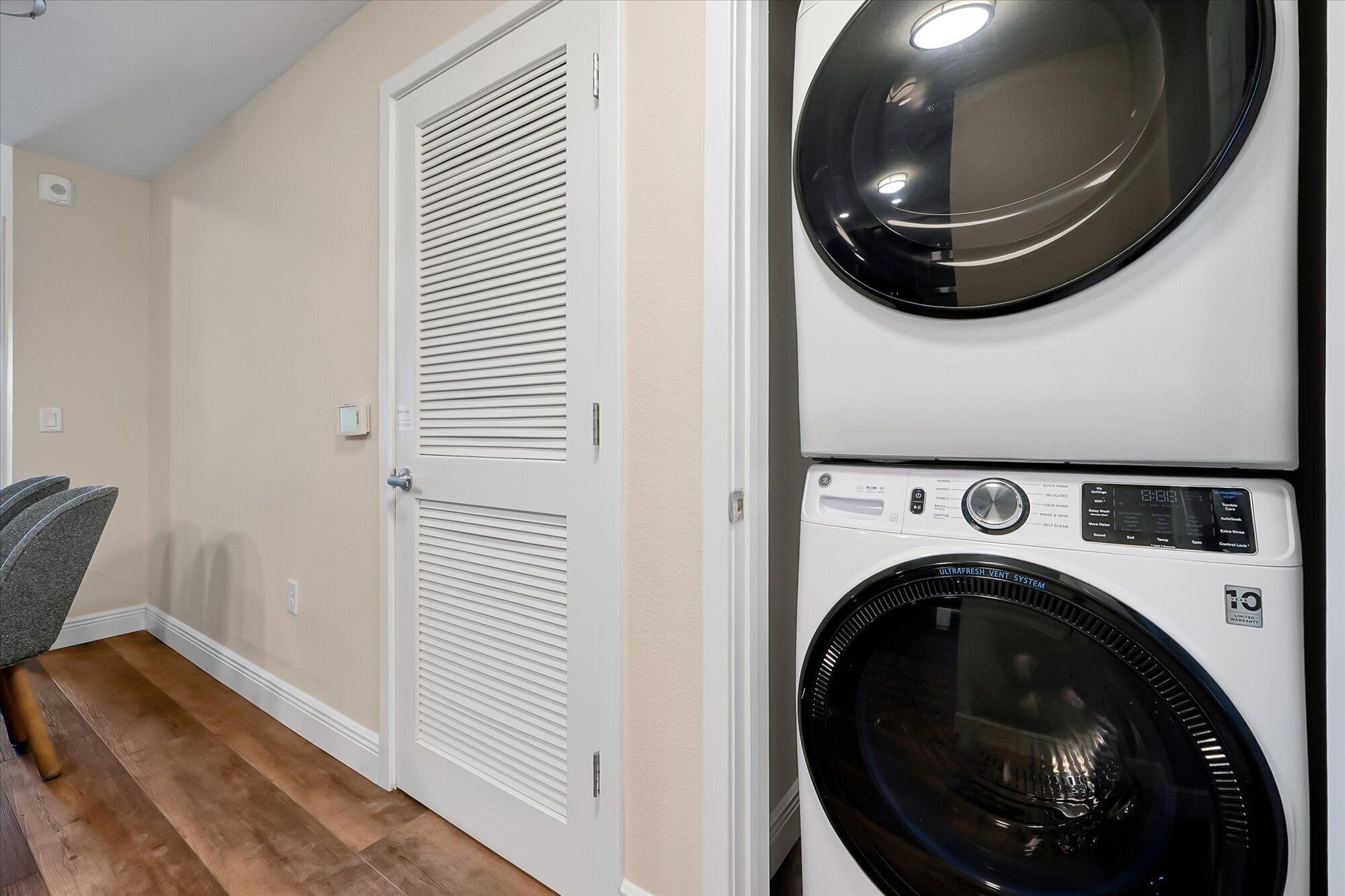 A laundry room with two stacked front-loading washing machines, a white door with a louvered design, and a wooden floor.