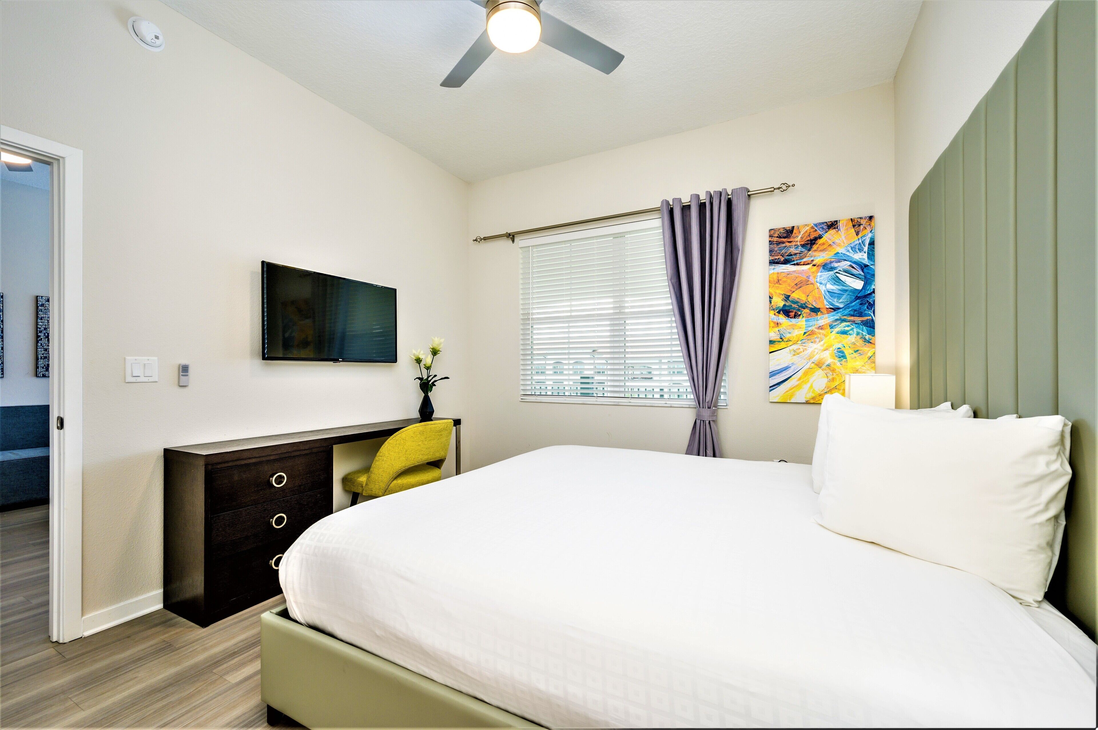 A bedroom with a bed, a desk, a chair, a television, a window with curtains, and a ceiling fan.