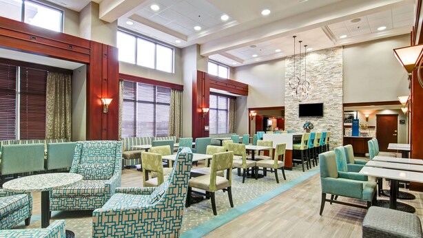 A spacious hotel lobby with multiple seating areas, including sofas and chairs, a central dining table, and a stone accent wall.
