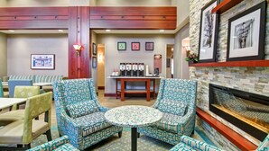 A lobby with patterned armchairs, a round table, a fireplace, framed artwork, and a coffee station.
