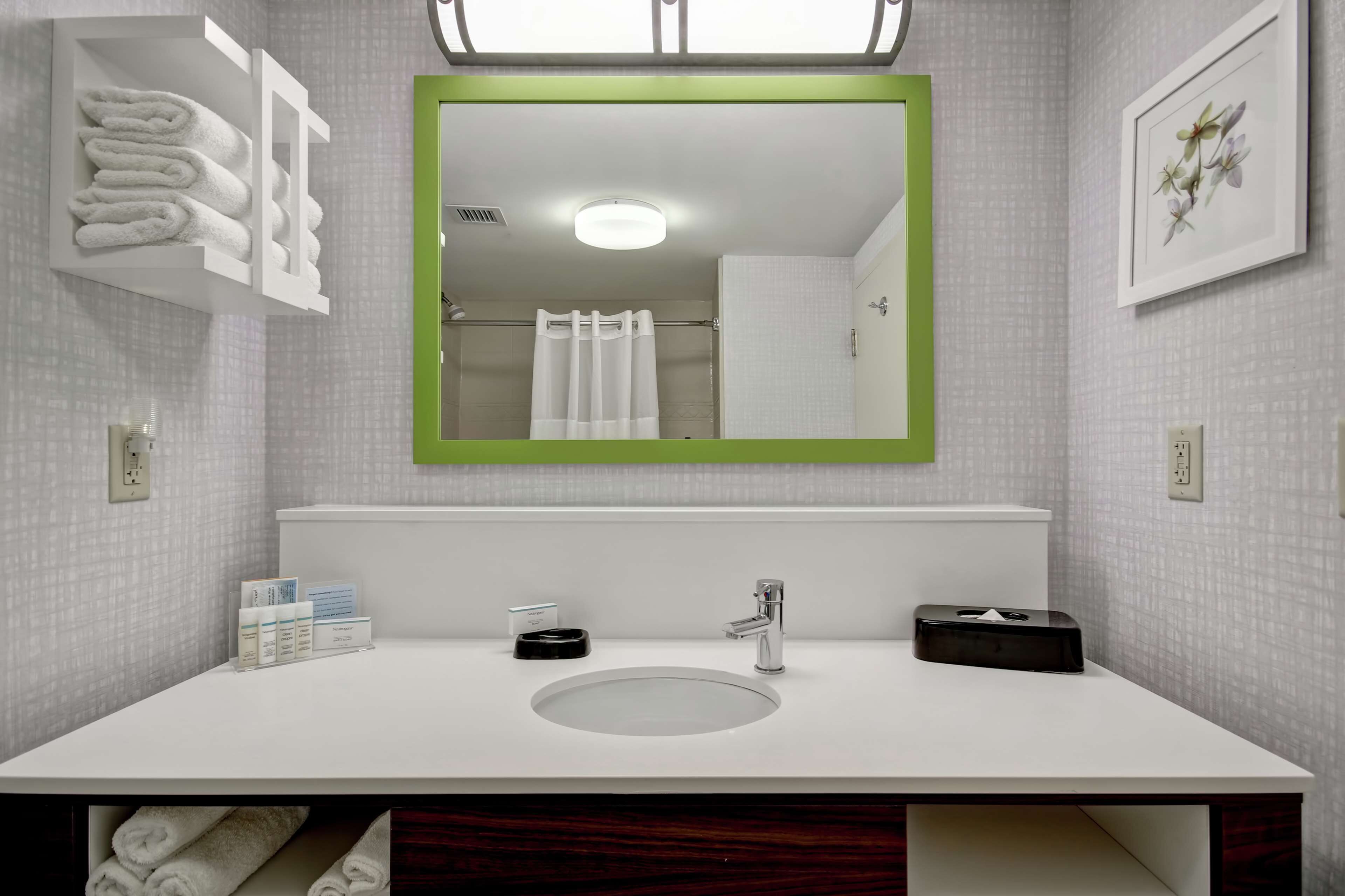 Un baño moderno con un lavamanos blanco, un espejo con marco verde y toallas ordenadas sobre una repisa.