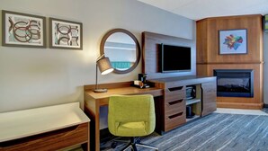 A modern hotel room with a wooden desk, a green chair, a flat-screen TV, a fireplace, and abstract wall art.