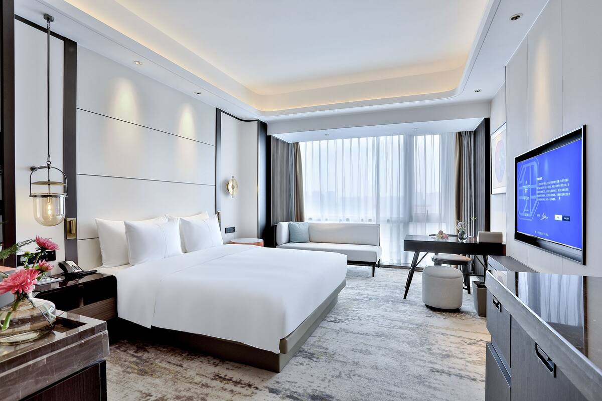 Un moderno cuarto de hotel con una cama grande, un escritorio, un televisor y vistas a la ciudad a través de amplias ventanas.