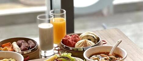 Un buffet de petit-déjeuner proposant divers plats, notamment des gaufres, des omelettes et des viennoiseries, servi sur une table en bois avec vue sur la rue.