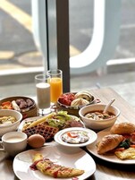 A breakfast buffet with various dishes, including waffles, omelets, and pastries, served on a wooden table with a view of the street outside.