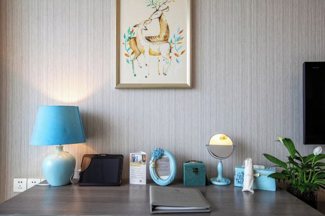A hotel room with a desk, a lamp, a framed artwork, a plant, and a television.
