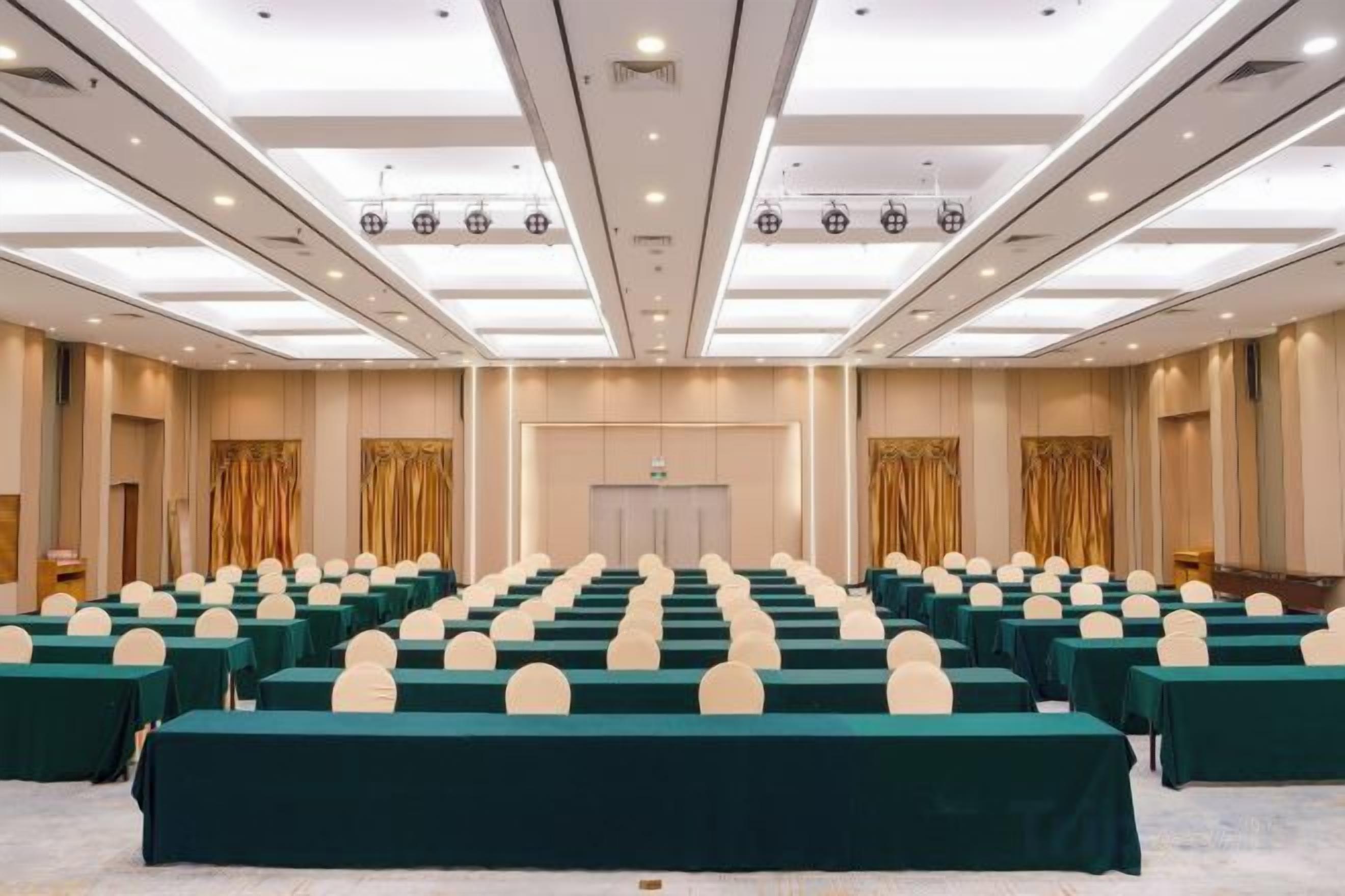 Sala de conferência com fileiras de mesas e cadeiras, um palanque e uma porta ao centro.