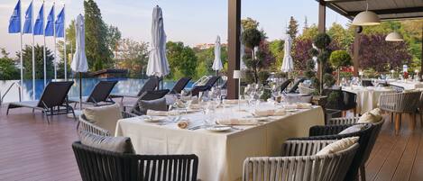 An outdoor dining area with tables set for a meal, surrounded by lounge chairs and umbrellas, overlooking a pool and greenery.
