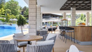 A covered outdoor seating area with a pool, wooden tables, and chairs, and a bar with a fruit display.