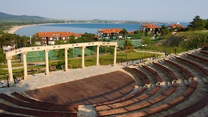 An outdoor amphitheater with tiered seating, a stone floor, and a view of a beach and buildings in the background.