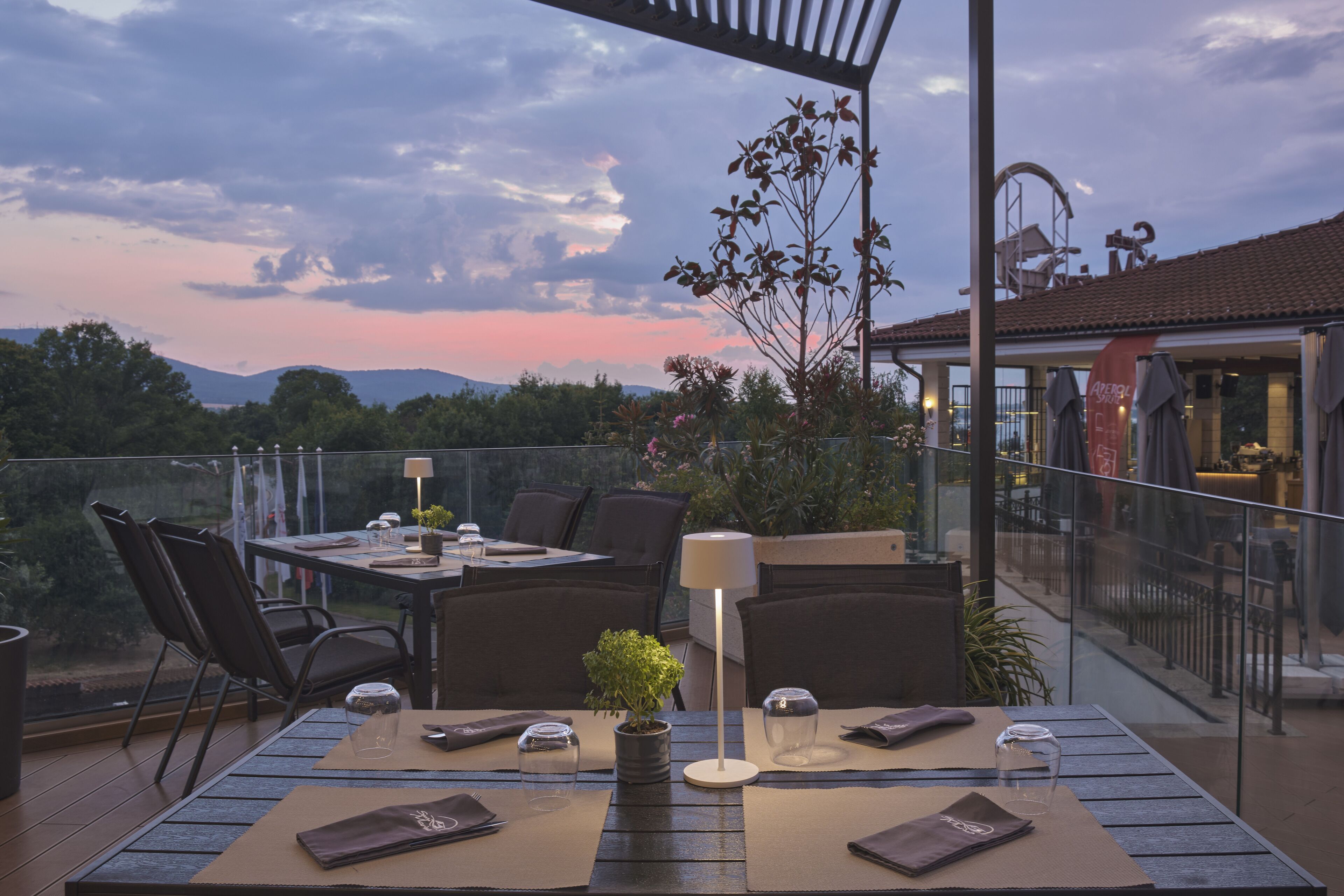 An outdoor dining area with a table set for two, surrounded by chairs, a potted plant, and a lamp.