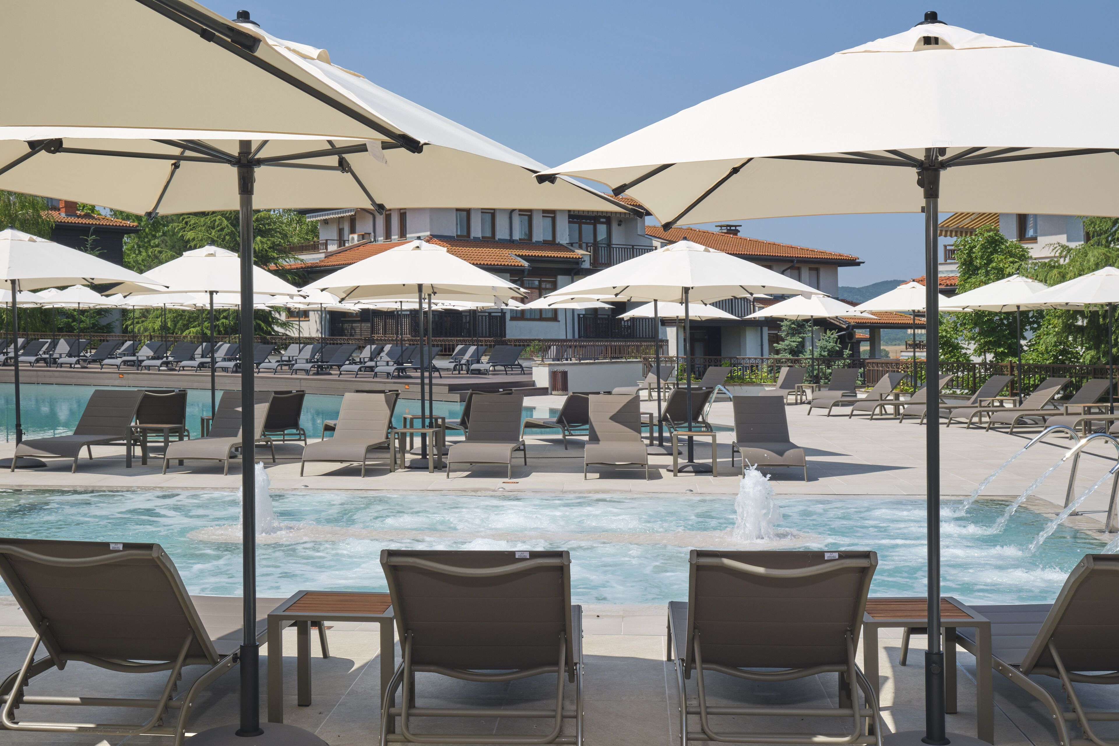 A poolside area with lounge chairs and umbrellas, a pool with a water feature, and a building in the background.