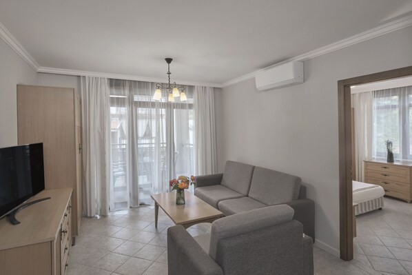 A modern living room with a sofa, a coffee table, a TV, and a window with sheer curtains. - Santa Marina Holiday Village (Sozopol)