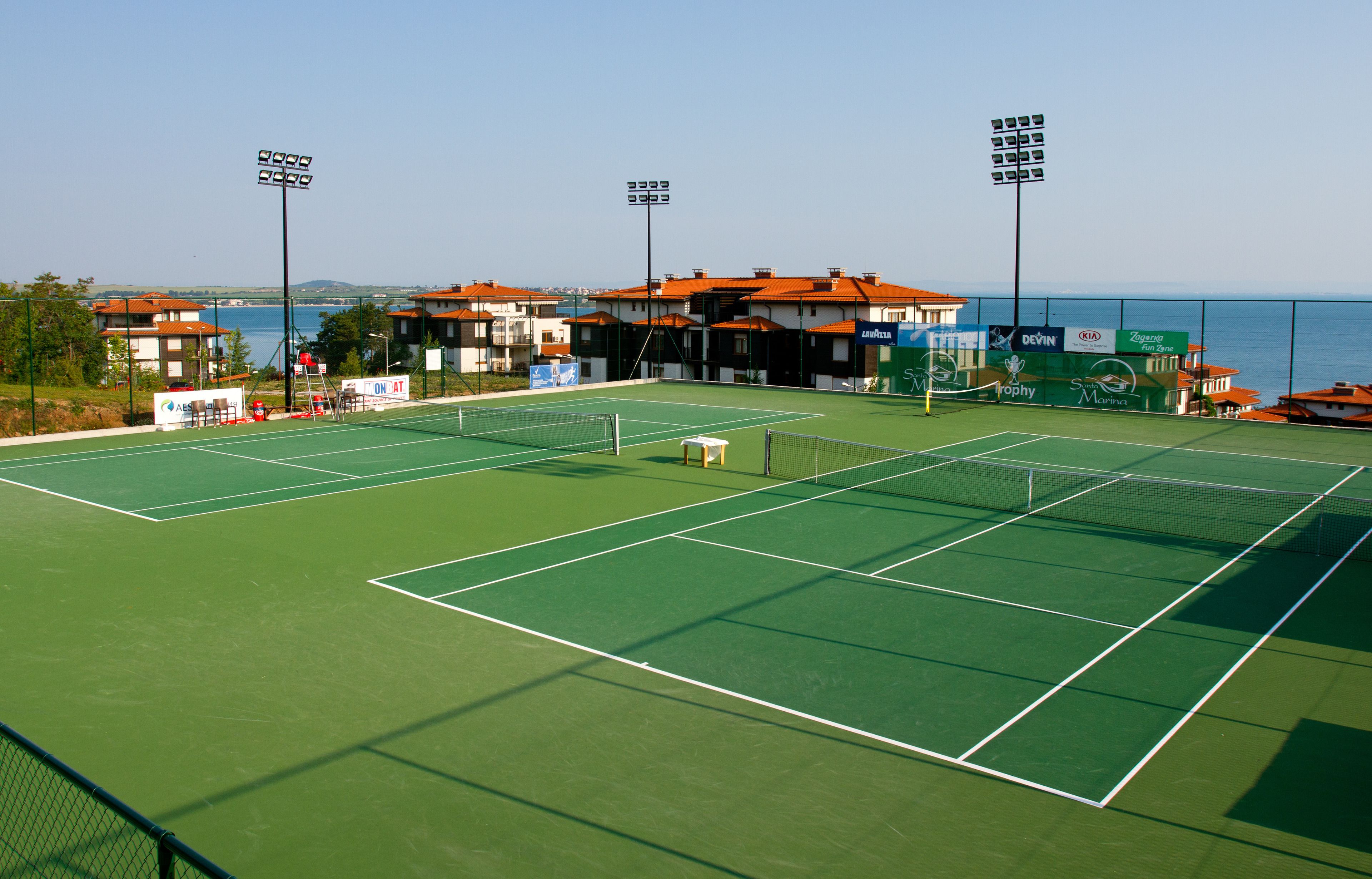 A tennis court with a net, surrounded by buildings with red roofs, and a clear view of the sea in the background.