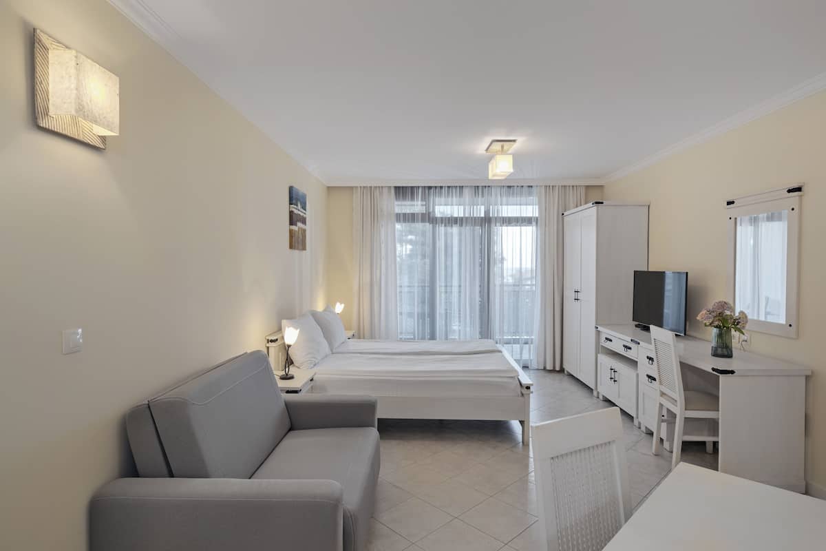 A modern hotel room with a sofa, a desk, a TV, and a large window with curtains.