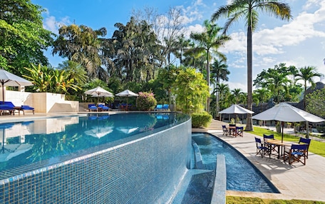 A clear blue infinity pool with lounge chairs and umbrellas, surrounded by lush tropical vegetation.