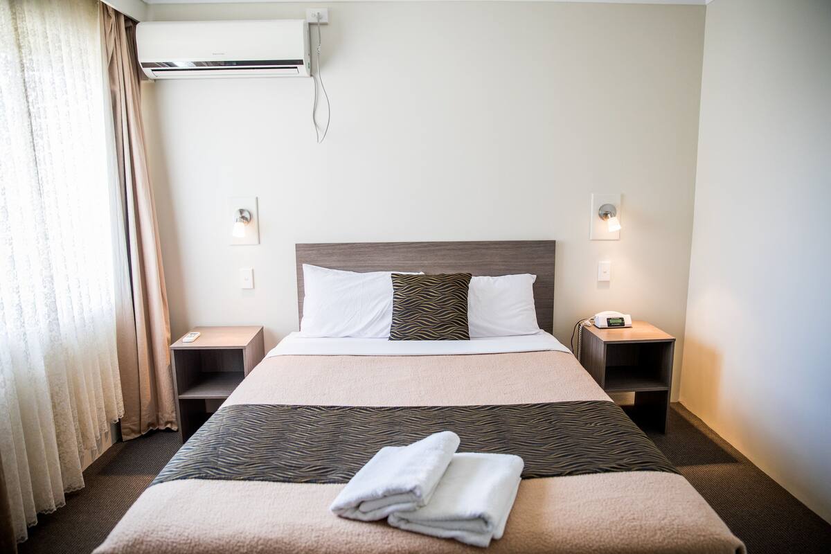 Habitación de hotel con una cama grande, dos mesitas de noche, un equipo de aire acondicionado y una ventana con cortinas.