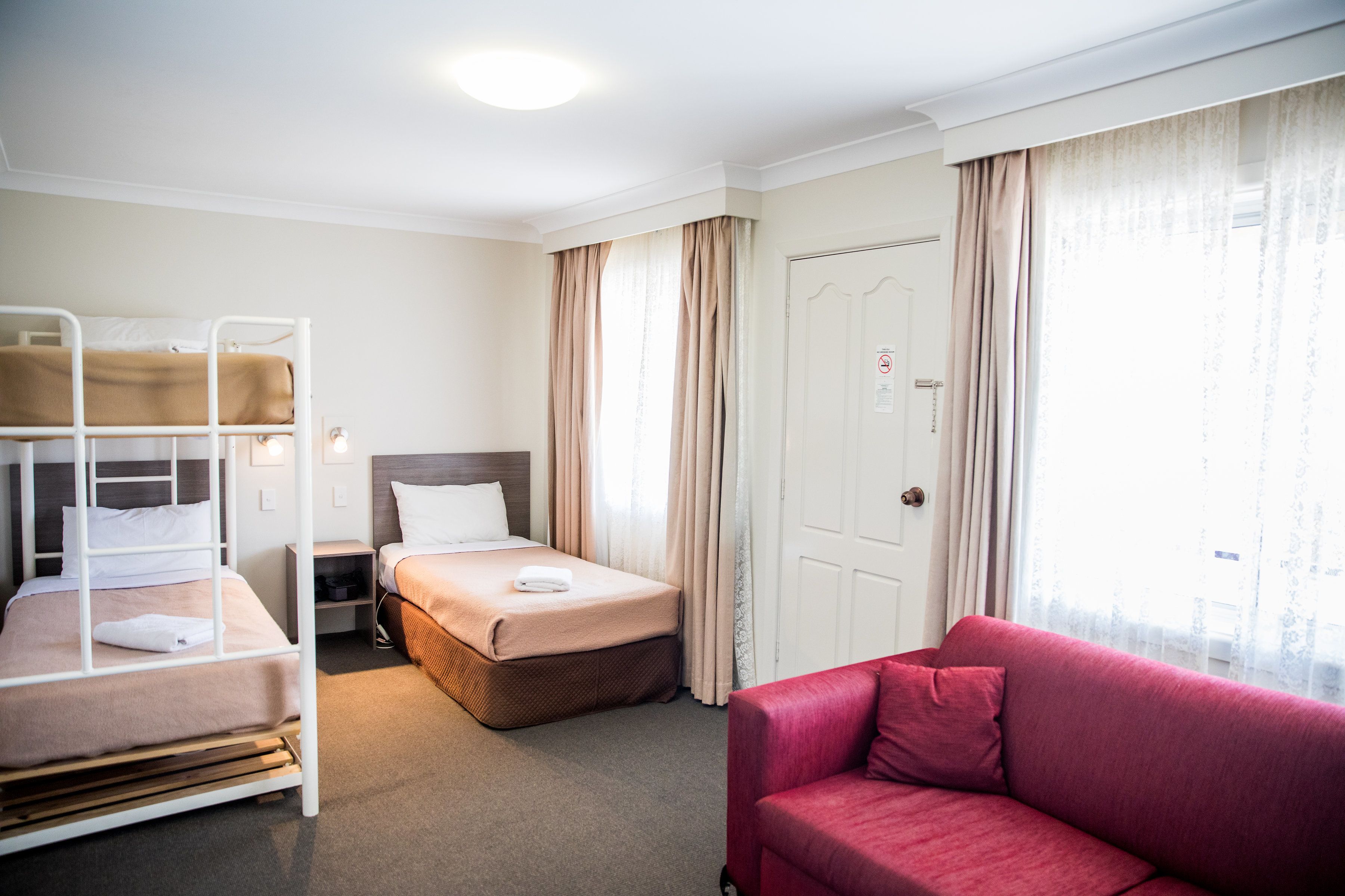 A hotel room with a bunk bed, a single bed, a red sofa, and a door with a sign.