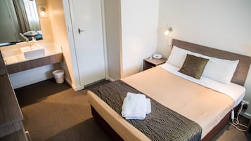 Una camera d'albergo con un letto, un comodino con una lampada, un bagno con lavandino e specchio, e una porta.