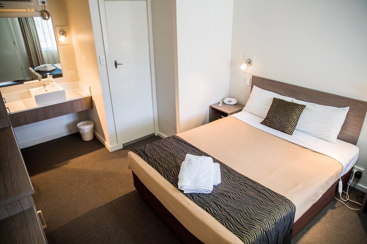 Habitación de hotel con cama, mesita de noche con lámpara, baño con lavamanos y espejo, y una puerta.