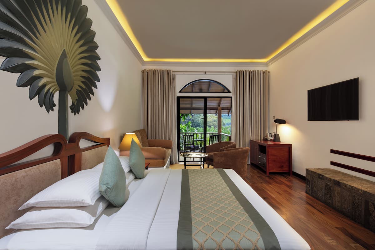 Habitación de hotel con una cama grande, una zona de estar con sofá y sillones, una mesita y un balcón con vistas a zonas verdes.