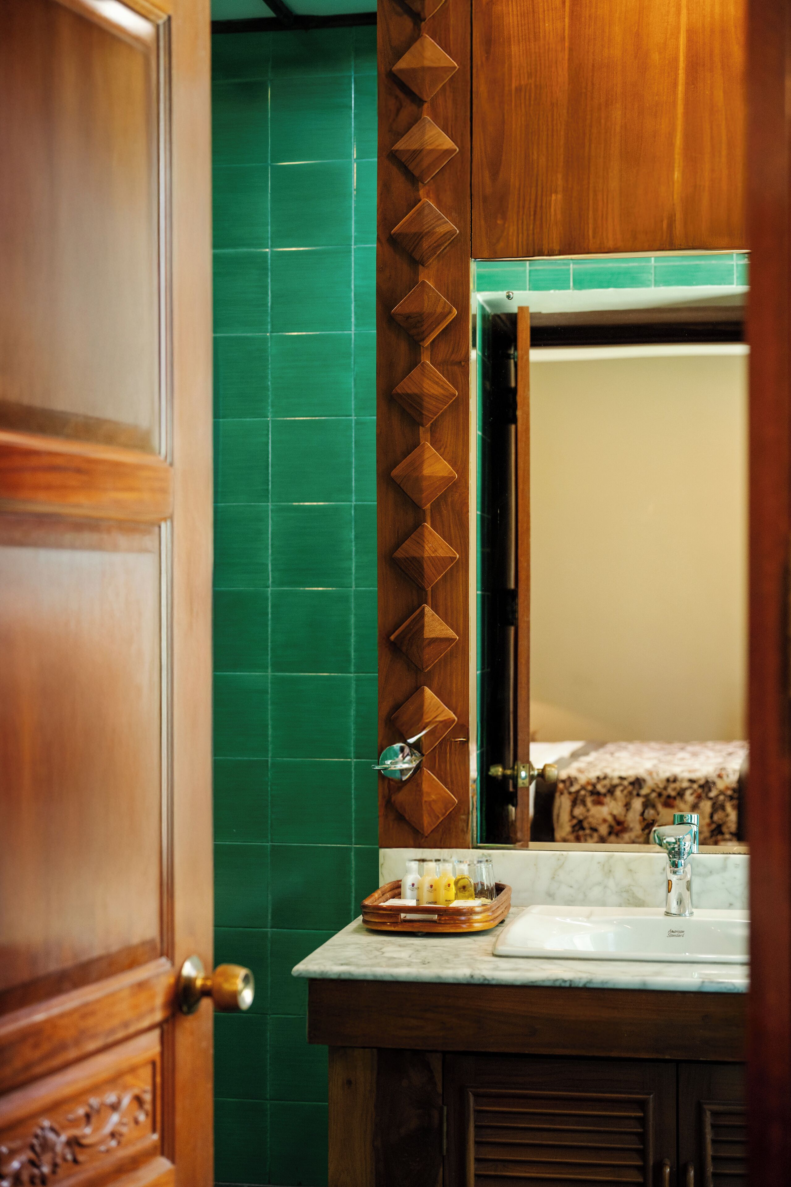 Een badkamer met een houten deur, groene betegelde wanden, een wastafel met spiegel en toiletartikelen op het aanrecht.