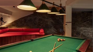 Una mesa de billar con bolas y tacos, un sofá rojo e iluminación colgante verde en una habitación con pared de piedra de fondo.