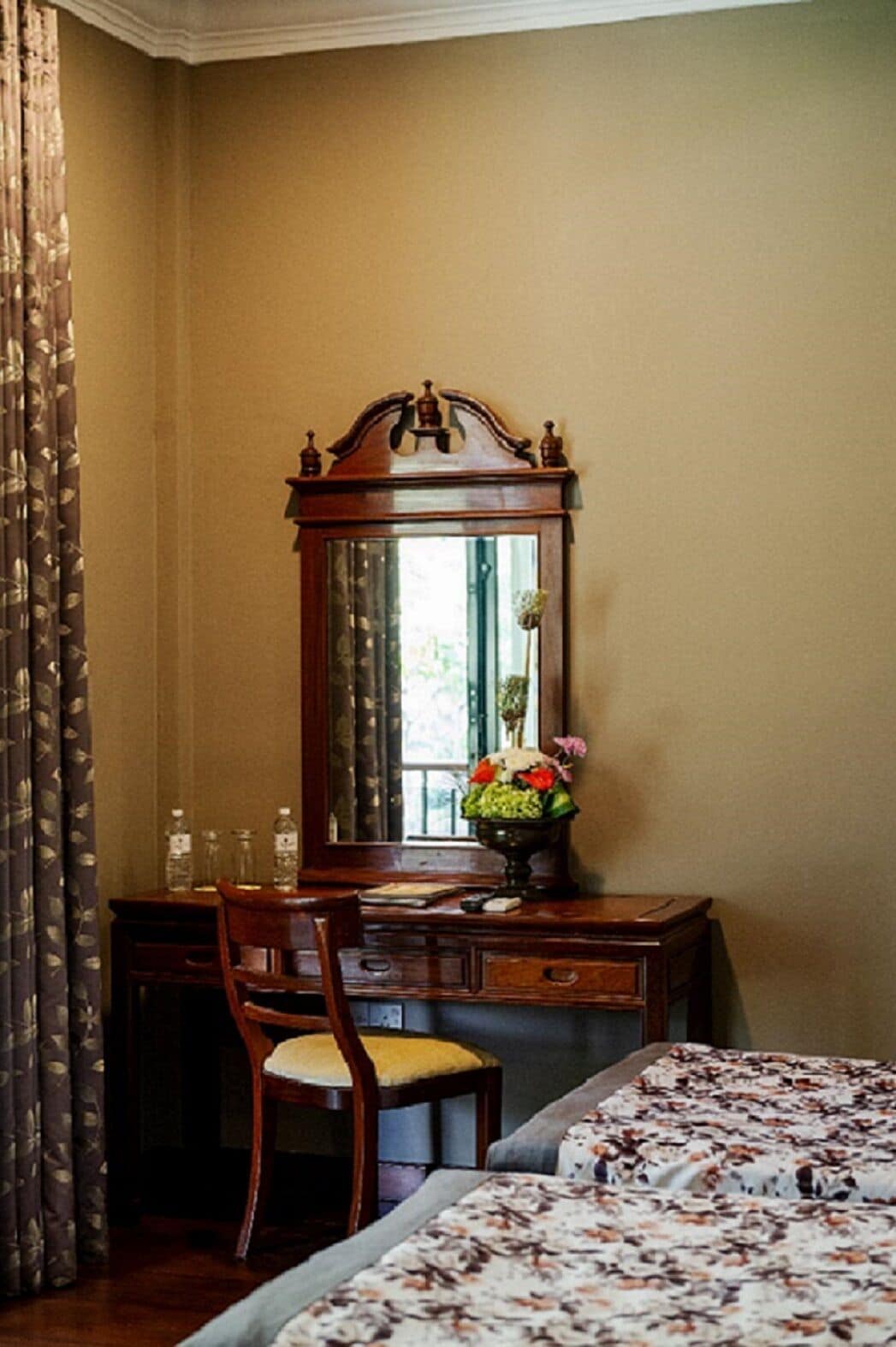 A bedroom with a wooden desk, a chair, a bed with a floral bedspread, a mirror, and a window with curtains.