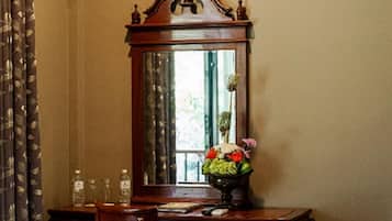 A bedroom with a wooden desk, a chair, a bed with a floral bedspread, a mirror, and a window with curtains.