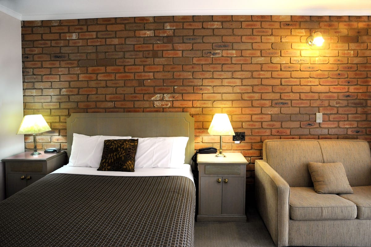 A hotel room with a brick wall, a bed with a patterned bedspread, two bedside tables with lamps, and a sofa.