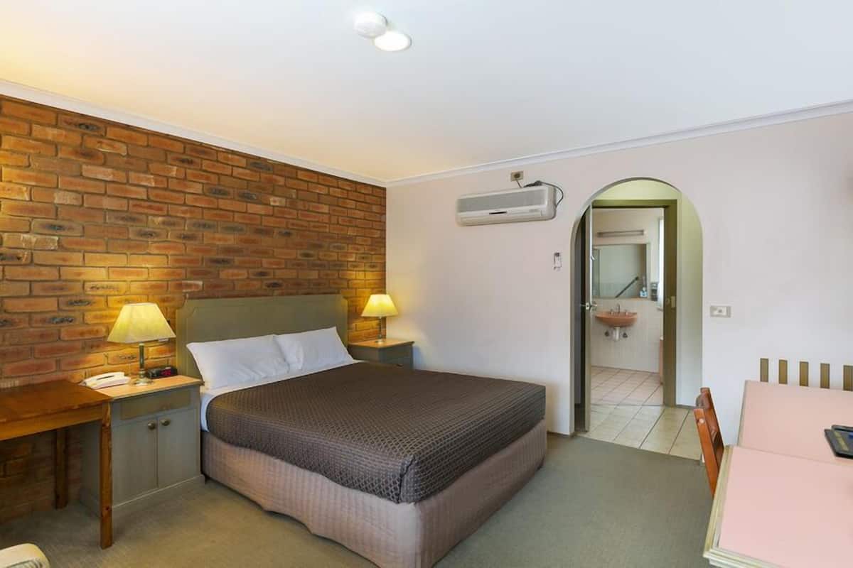 A bedroom with a brick wall, a bed with a brown blanket, two bedside lamps, a wooden nightstand, a small table, and an air conditioning unit.