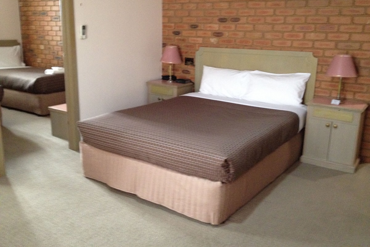 A hotel room with a brick wall, a bed with a brown bedspread, two bedside tables with lamps, and a nightstand with a lamp.