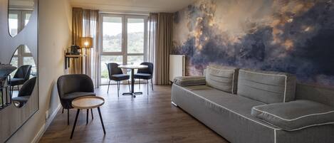 Een moderne woonkamer met een bank, stoelen, een klein rond tafeltje en een groot raam met gordijnen.