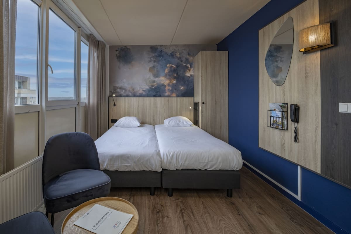 A hotel room with two beds, a chair, a round table with a menu, and a large window.