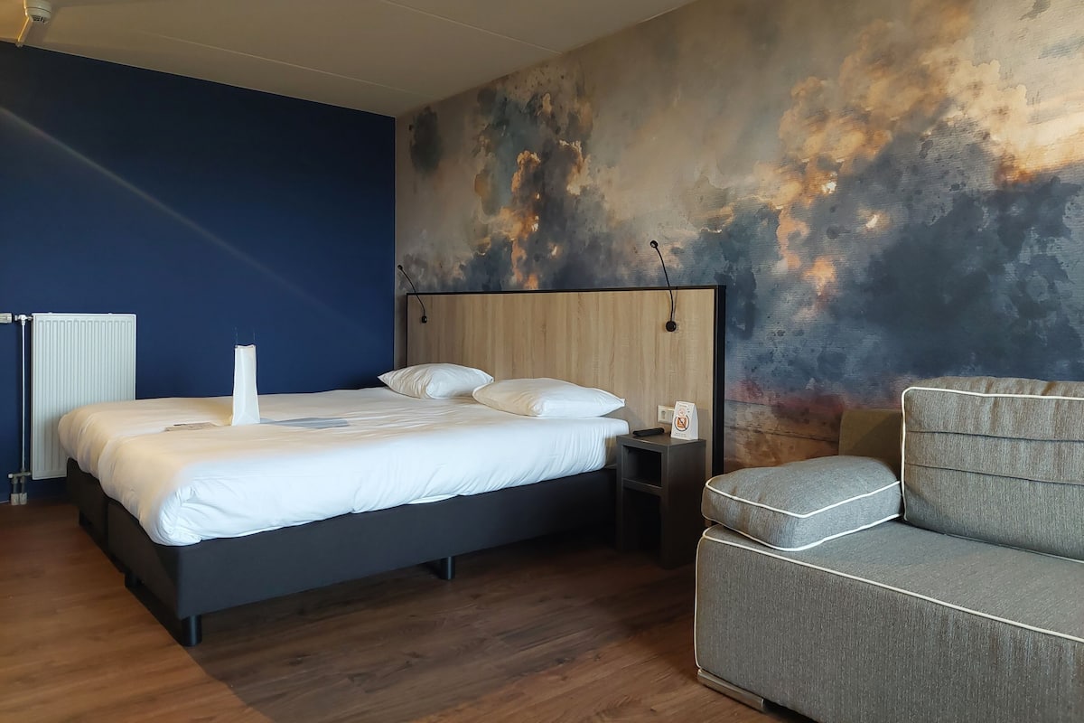 A modern hotel room with a large bed, a grey sofa, a wooden headboard, and a textured wall mural.