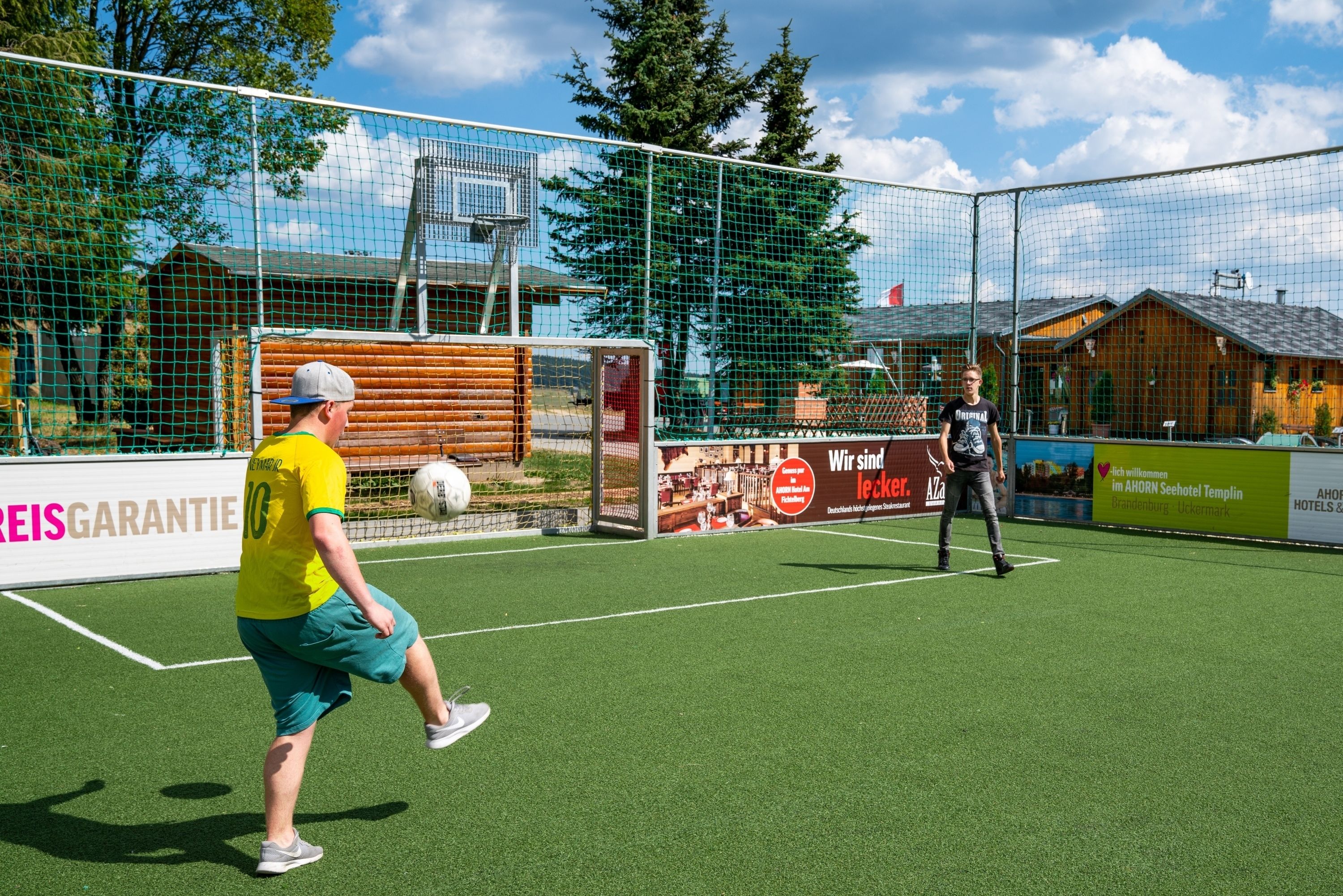 A soccer field with a goal net, a player in a yellow jersey, and another player in a black jersey.