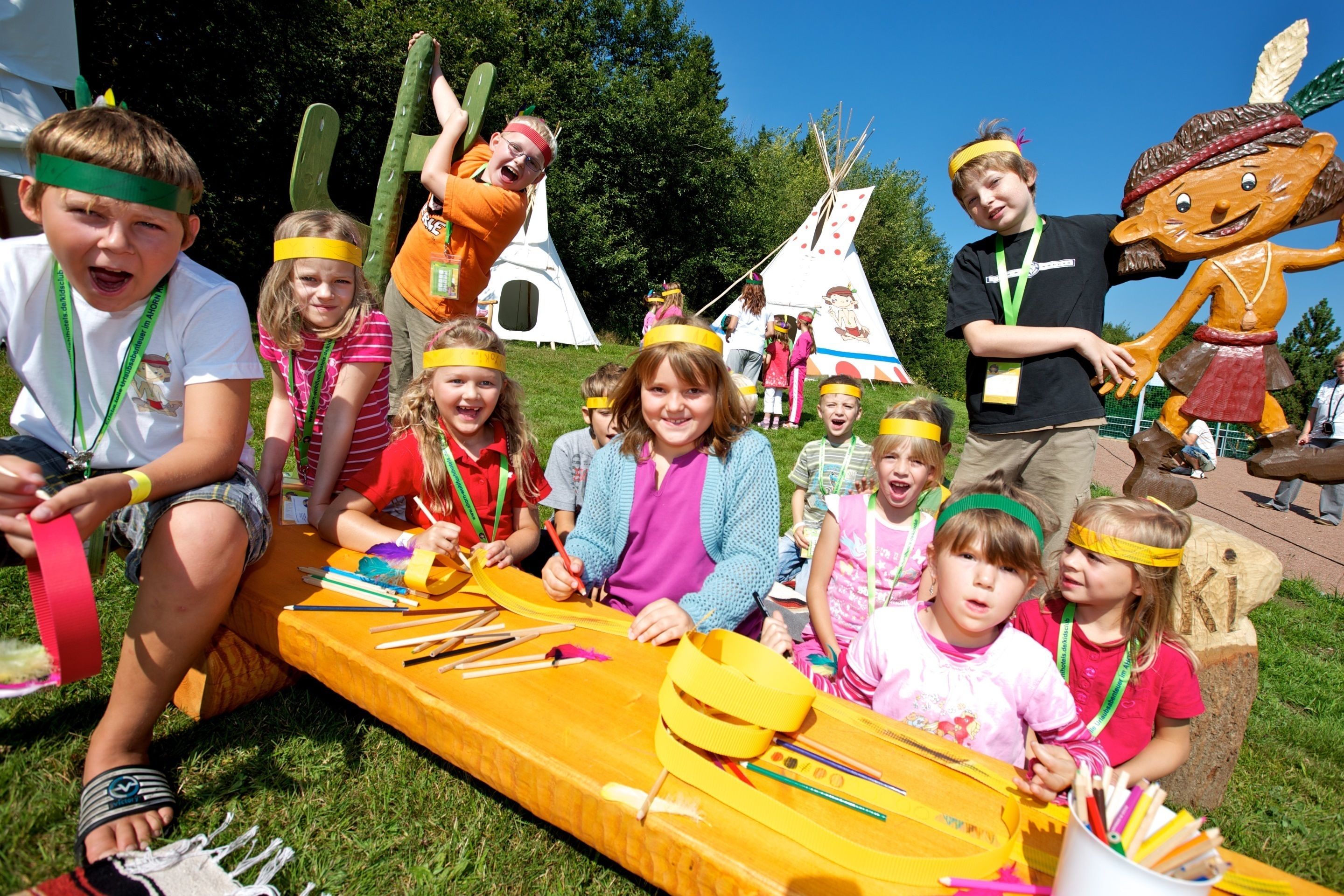 A group of children engaged in an outdoor activity with a long yellow table, various tools, and a large mascot figure.