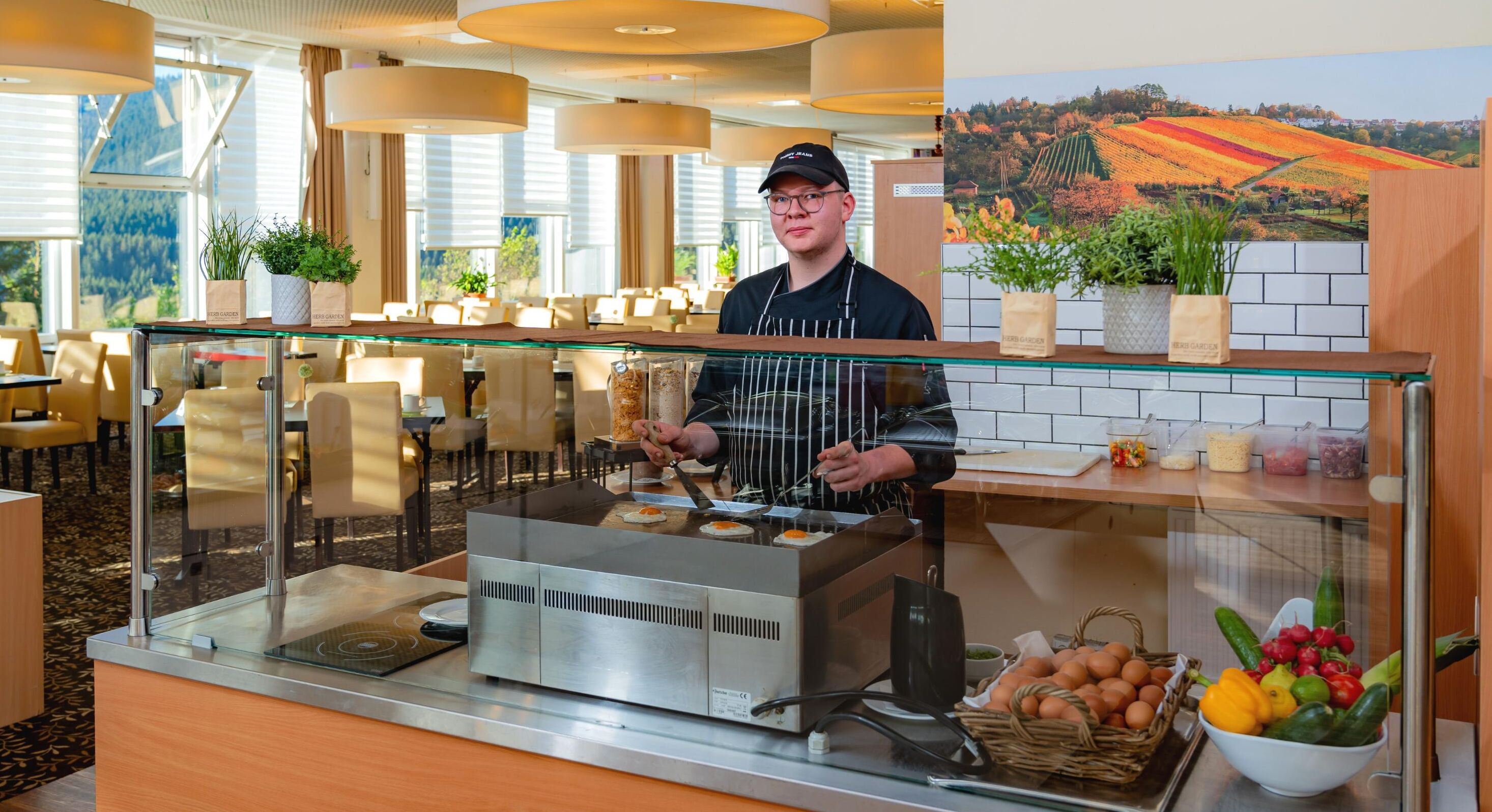Estación de desayuno con un chef preparando alimentos, un expositor de vidrio y una variedad de frutas y verduras frescas.