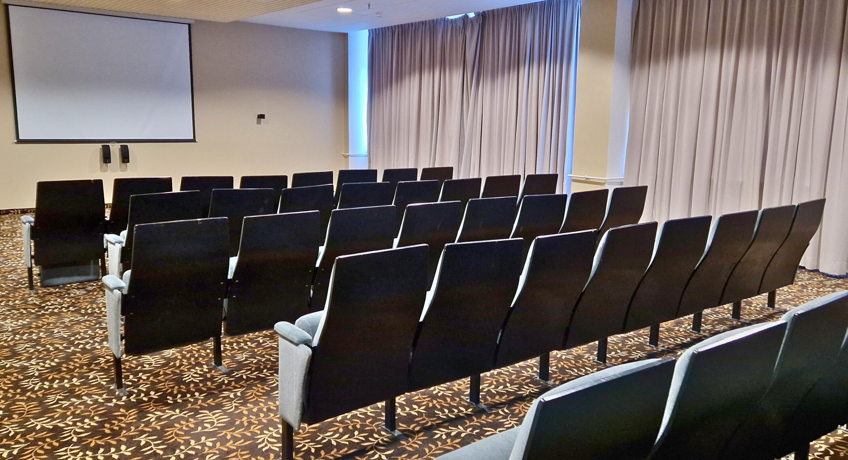 A conference room with rows of chairs, a projector screen, and curtains.