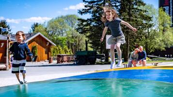 A child jumping on a trampoline in a park with wooden cabins in the background.