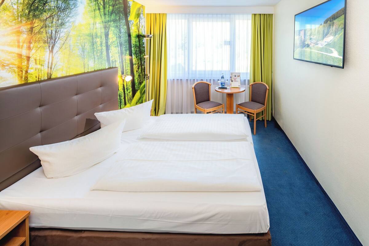 Une chambre d’hôtel avec un lit, un bureau, deux chaises, une fenêtre avec des rideaux et un tableau au mur.